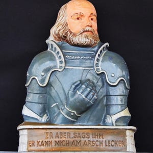 Puede incluir: Un busto de cerámica de un hombre con armadura, barba y bigote. El busto está pintado en tonos azules, blancos y marrones. El busto está montado sobre una base con el texto "ER ABER, SAGS IHM ER KANN MICH AM ARSCH LECKEN" en alemán.