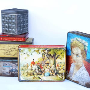 May include: A collection of vintage metal tins in various shapes and sizes. The tins feature decorative designs, including a portrait, a hunting scene, and geometric patterns. Some tins have text, such as "Cotillon Cigarettes" and "Pfeiffer Kaffee Ersatz".