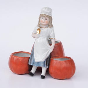 Puede incluir: Figura de cerámica vintage de una niña con gorro y delantal de chef, sosteniendo una cuchara. La figura está colocada frente a tres recipientes naranjas con forma de calabaza. El atuendo de la niña tiene ribetes dorados.