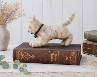 May include: A needle-felted cat figurine with cream and grey tabby stripes, featuring a dark brown collar with a small metal bell. The cat is posed in a standing position on top of a large, rectangular, dark brown antique book with gold lettering.