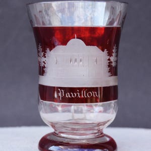 May include: A clear glass with a red rim and base, featuring an etched design of a building and trees. The word "Pavillon" is etched in a band around the glass. The glass has a flared shape and a rounded base.