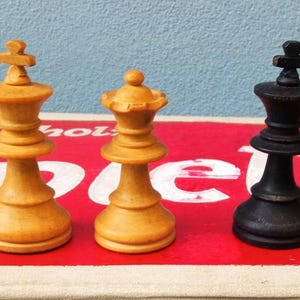 Puede incluir: Cuatro piezas de ajedrez: dos de color claro y dos negras. Las piezas claras son un rey y una reina, mientras que las piezas negras también son un rey y una reina. Las piezas están sobre una superficie roja con texto blanco.