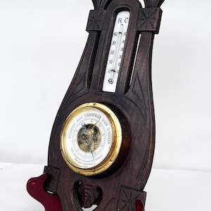 May include: Antique wooden barometer and thermometer combination. The dark wood frame features ornate carvings and a decorative floral element at the top. The thermometer displays temperature in Celsius, and the barometer has a brass-colored face.