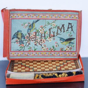 Puede incluir: Caja roja vintage con un juego de ajedrez. La tapa presenta un fondo azul claro con ilustraciones de pájaros, flores y la palabra "JUMA". En el interior, se ve un pequeño tablero de ajedrez y piezas de ajedrez. La caja está abierta.