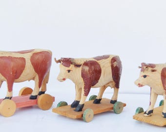 Op de afbeelding: Drie vintage houten speelgoedkoeien met rode en bruine markeringen, gemonteerd op houten platforms met wielen. De koeien zijn geschilderd in tinten beige, rood en bruin, en de wielen zijn in verschillende kleuren geschilderd.