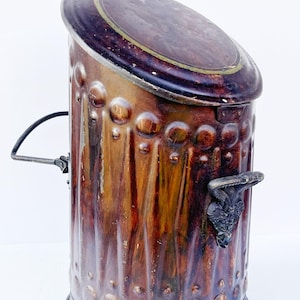 Könnte beinhalten: Ein antiker, zylindrischer Metallbehälter mit einem Klappdeckel. Der Behälter ist hauptsächlich braun mit einer strukturierten Oberfläche und dekorativen kreisförmigen Akzenten. Ein dunkler Griff und ein verzierter Verschluss sind sichtbar.