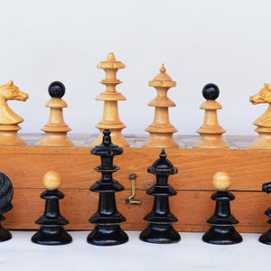 Puede incluir: Un juego de ajedrez vintage con piezas negras y crema. Las piezas están dispuestas sobre un tablero de madera dentro de una caja de madera. Las piezas de ajedrez incluyen caballos, torres, peones, una reina y un rey. El juego se exhibe sobre una superficie blanca.