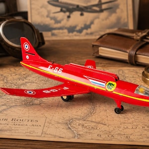 Pode incluir: Um avião de brinquedo de metal vermelho brilhante com listras amarelas, uma cabine transparente e trem de pouso preto. A fuselagem está marcada com o texto E-G6 e F-G6, juntamente com cocardas circulares azul, branca e vermelha e um emblema.