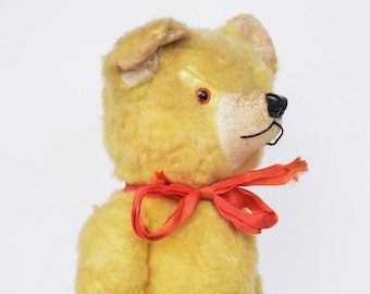 Puede incluir: Un oso de peluche de estilo vintage con pelaje dorado, nariz negra y ojos rojos. El oso tiene un hocico color crema y lleva una cinta roja alrededor del cuello. El juguete de peluche es un artículo clásico para niños.
