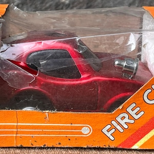 Könnte beinhalten: Ein rotes Spielzeugauto vom Typ "Fire Chief" in seiner originalen orangefarbenen Pappschachtel. Das Auto ist durch ein transparentes Kunststofffenster sichtbar. Auf der Schachtel steht "FIRE CHIEF" in weißer Schrift.