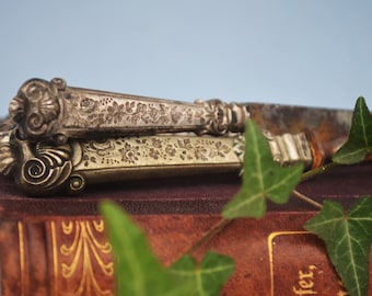 May include: Two antique silver knives with ornate floral designs on the handles, resting on a brown leather-bound book with gold accents and green ivy leaves.