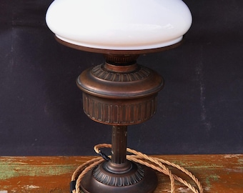 May include: An antique table lamp with a white glass shade and a bronze-colored metal base. The shade has a ruffled edge. The lamp has a decorative base and a braided cord with a white plug. The lamp is on a wooden surface.