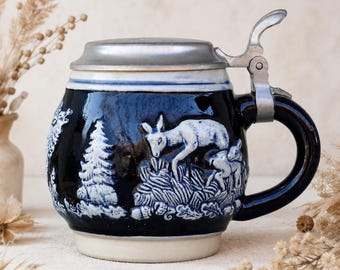 Puede incluir: Una jarra de cerveza de cerámica azul oscuro con un relieve blanco que muestra un ciervo en un entorno forestal. La jarra tiene una base clara, un asa curva y una tapa metálica con bisagras y una palanca en el borde superior.