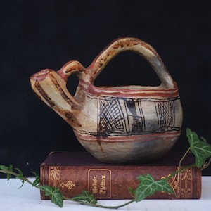 Pode incluir: Jarro de cerâmica artesanal, em estilo antigo, com asa e bico. Apresenta uma base bege com detalhes pintados em vermelho e preto, incluindo um padrão geométrico. Descansa sobre um livro vintage com capa escura, com hera em primeiro plano.