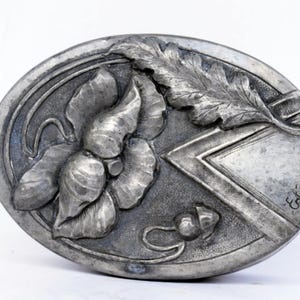 May include: Oval, silver-toned metal decorative piece with a floral and leaf design. The design includes a large flower, a leaf, and a small bud. The piece has an Art Nouveau style.