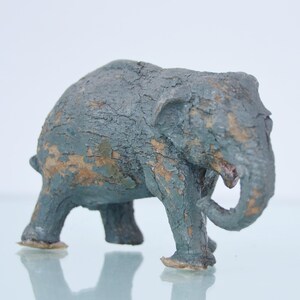 Peut inclure: Petite figurine d'éléphant patinée, dans des tons de bleu et de brun. La sculpture présente une surface texturée, avec des signes d'usure visibles, lui donnant un aspect vintage. L'éléphant est posé sur une surface réfléchissante.