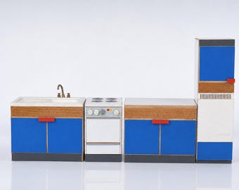 May include: A miniature toy kitchen set featuring a sink unit, a stove with four burners, a counter cabinet, and a tall refrigerator. The pieces have blue cabinet doors, white surfaces, wood-grain accents, and small red handles on a gray base.