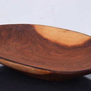 May include: A large, oval-shaped wooden serving bowl with dark brown wood grain and two dark brown handles. The bowl is made of natural wood and has a rustic, handcrafted look.