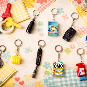 May include: A collection of novelty keychains in various shapes and colors, including keys, a camera, a miniature detergent box, and a miniature gas pump. The keychains are arranged on a patterned surface.