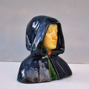 Pode incluir: Um busto de cerâmica de uma pessoa vestindo uma capa com capuz azul com detalhes verdes e laranja. O rosto da pessoa é visível e tem uma tez amarela.
