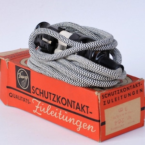 Op de afbeelding: Een opgerold zwart-wit elektrisch snoer met een stekker, rustend op een vintage oranje doos. De doos heeft Duitse tekst en een logo. Het snoer is voorzien van een zigzagpatroon.