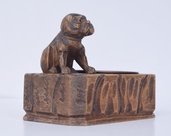 May include: A wooden sculpture of a dog sitting atop a rectangular box. The dog is brown with detailed carving, and the box has a textured, carved design. The piece is likely a decorative item or a small box.