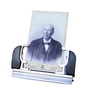 May include: A black and silver photo holder with a vintage black and white photo of a man with a mustache and beard. The photo is in a silver frame with a black border. The photo holder has a silver bar at the bottom with the text "Kronstadt, Brassau" on it.