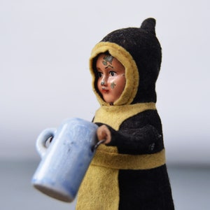 Puede incluir: Una muñeca vintage que lleva un abrigo negro con capucha y ribete amarillo y sostiene una jarra de cerámica azul.