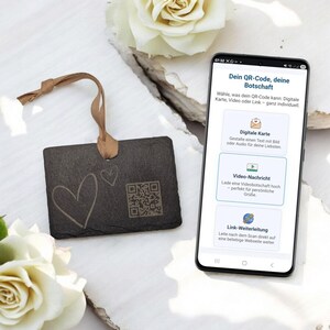 Könnte beinhalten: Ein schwarzer rechteckiger Schiefer mit einem QR-Code und zwei Herzdesigns, mit einem braunen Band befestigt. Ein Smartphone zeigt Optionen für digitale Karten, Videobotschaften und Website-Links. Der Text auf dem Telefon lautet "Dein QR-Code, deine Botschaft."