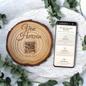 Könnte beinhalten: Ein Holzscheibe mit den Worten "Von Herzen" und einem QR-Code. Das Wort "Scan" befindet sich unter dem QR-Code. Ein Smartphone zeigt eine Webseite mit Optionen für digitale Karten und Videonachrichten.