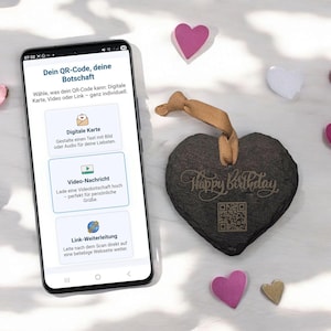 Könnte beinhalten: Ein Smartphone zeigt eine QR-Code-Nachricht auf Deutsch mit Optionen für digitale Karten, Videobotschaften und Links. Ein herzförmiger Schieferornament mit "Happy Birthday" und einem QR-Code hängt daneben, umgeben von rosa und goldenen Herzdekorationen.