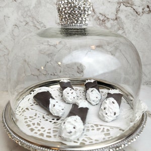 May include: A silver serving tray with a glass dome, holding several cannoli pastries. The cannoli have dark chocolate shells and white, creamy filling. The dome is topped with a sparkling, crystal-like handle.