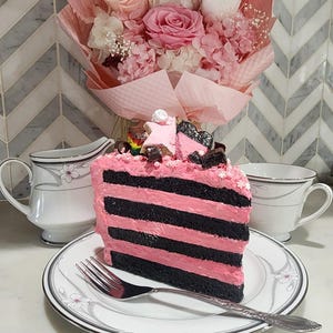 May include: A slice of layered cake with pink frosting and black stripes, topped with star-shaped decorations, sits on a plate with a fork. A bouquet of pink roses and other flowers is in the background, along with a creamer and teacup.
