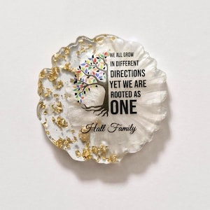 Könnte beinhalten: Ein Untersetzer aus Kunstharz mit weißer Basis und Goldflocken. Der Untersetzer zeigt eine Baumgrafik mit bunten Blättern und dem Text "WE ALL GROW IN DIFFERENT DIRECTIONS YET WE ARE ROOTED AS ONE Hall Family."