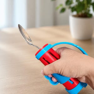 May include: A silver-colored metal spoon attached to a cylindrical red and blue plastic adaptive grip handle. The handle features a blue curved strap for securing the hand and a molded blue finger-grip section on the underside for stability.