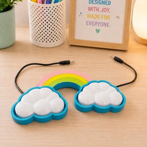 May include: A rainbow-shaped assistive switch featuring two white cloud-shaped buttons with blue borders. The device includes two black cables with 3.5mm jacks extending from the sides. The rainbow arch is coloured in pink, yellow, and green stripes.