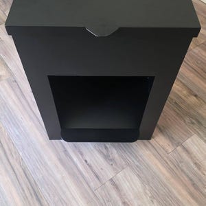Puede incluir: Un contenedor de almacenamiento rectangular de color negro mate con una tapa abatible que cuenta con dos bisagras metálicas en el borde superior. El frente tiene un gran compartimento rectangular abierto y una pequeña pestaña en la tapa.