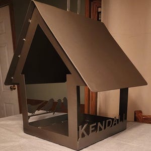May include: A dark brown metal bird feeder shaped like a house with a peaked roof. The structure features a cutout silhouette of three birds perched on a branch along the back panel and the name KENDALL cut out in capital letters on the front edge.