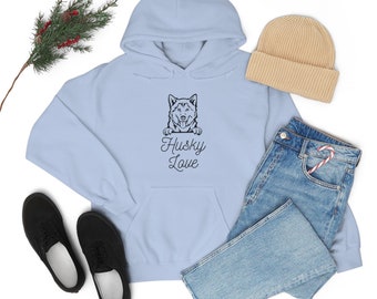 Op de afbeelding: Een lichtblauwe hoodie met een zwarte husky-hond die over de bovenkant kijkt en de tekst "Husky Love" eronder. De hoodie ligt op een wit oppervlak met een paar zwarte schoenen, een blauwe jeans en een beige muts.