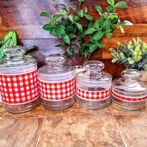 May include: Four clear glass jars with red and white gingham ribbon around the top. The jars are arranged in a row from largest to smallest.