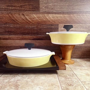 May include: Two vintage yellow casserole dishes with white lids and black handles. One dish sits on a wooden stand, the other on a black tray with wooden handles. The background is a wood-paneled wall.