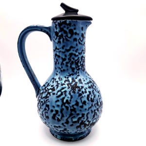 May include: A blue ceramic pitcher with a black lid and handle. The pitcher has a textured surface with a speckled pattern of black spots. The handle is curved, and the lid has a small spout.