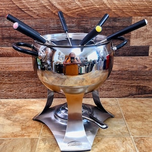 May include: A stainless steel fondue pot with black handles and forks. The pot has a reflective surface, and the base is made of brushed metal. The fondue forks have colored tips. The brand name "Trudeau" is visible on the base.