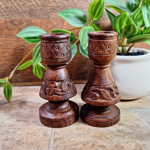 May include: Two dark brown wooden candle holders with intricate carvings. The holders are made of a dark wood with a natural finish. The holders are both in the shape of a vase with a wide base and a narrow neck.