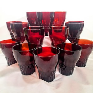 May include: A collection of ruby red drinking glasses. The glasses have a unique shape with a wide top and a textured, geometric pattern near the base. The glasses are arranged on a white surface, creating a striking display of glassware.