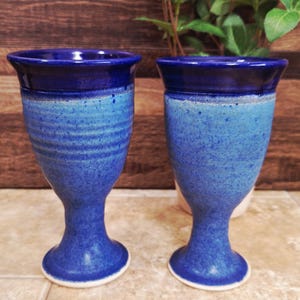 May include: Two handmade ceramic goblets with a blue glaze. The goblets have a dark blue rim, a lighter blue body, and a footed base. The textured surface adds to the rustic charm of the drinkware.