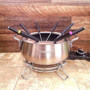 May include: A stainless steel Cuisinart fondue pot with a chrome stand and black handles. The pot includes six fondue forks with colored tips. The electric base has a temperature control dial.