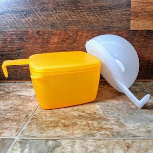 May include: A yellow plastic food storage container with a lid and a handle. The container is sitting on a brown tiled surface. A white plastic container with a lid is also in the image.