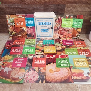 May include: A collection of vintage cookbooks with colorful covers, including titles like "Meat," "Dairy," "Dessert," and "Salads." A white box with "Cookbooks" printed on it sits in the center. The books are arranged on a wooden surface.