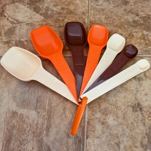 May include: A set of six measuring spoons in various colors, including orange, brown, and cream. The spoons are arranged in a fan shape, with measurements clearly marked on the handles. The spoons are made of plastic.